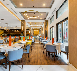 Long view over all view of restaurant with colorful blues, yellows and oranges. White marble and wood floors through out. Kitchen can be seen to far left corner and pizza over straight ahead.