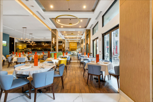 Long view over all view of restaurant with colorful blues, yellows and oranges. White marble and wood floors through out. Kitchen can be seen to far left corner and pizza over straight ahead.