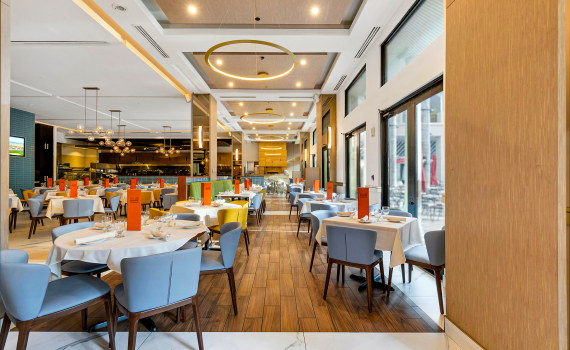 Long view over all view of restaurant with colorful blues, yellows and oranges. White marble and wood floors through out. Kitchen can be seen to far left corner and pizza over straight ahead.